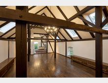 Innenraum eines historischen Dachgeschosses mit sichtbaren dunklen Holzbalken, schrägen Decken und mehreren Dachfenstern, die Tageslicht hereinlassen. Der Raum hat einen Holzboden, weiße Wände und eine zentrale, hängende Deckenleuchte.