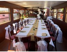 Ein festlich dekorierter Innenraum eines Bootes mit einer langen Tafel, die mit weißer Tischdecke, Jutestoff und kleinen Pflanzen in Gläsern geschmückt ist. Die Stühle sind mit weißem Organza geschmückt, und im Hintergrund ist eine kleine Bar sowie Fenster mit Blick auf das Wasser zu sehen.