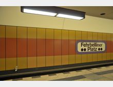 U-Bahn-Station mit gelb-orange verkleideter Wand und einem Schild mit der Aufschrift „Fehrbelliner Platz“. Der Boden besteht aus beigen Fliesen, und eine Leuchtstofflampe hängt an der Decke.