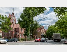 Straßenszene mit historischen Backsteingebäuden im Stil der Gründerzeit, umgeben von Bäumen. Autos parken am Straßenrand, während andere auf der Straße fahren; im Vordergrund führt eine Kreuzung durch das Bild.