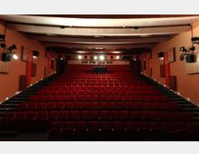 Innenraum eines Kinos mit mehreren Reihen roter gepolsterter Stühle, die in Stufen ansteigend angeordnet sind. Die Wände sind beige-orange gehalten und mit Holz- und Akustikelementen sowie Beleuchtungs- und Lautsprecherausrüstung ausgestattet.
