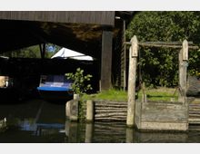 Ein hölzerner Unterstand mit einem Boot im Schatten, daneben ein rustikaler Holzziehbrunnen am Ufer eines kleinen Gewässers, umgeben von dichtem Grün und Grasflächen.