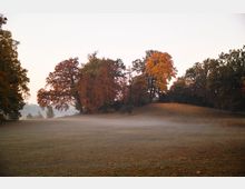 Weite, leicht nebelige Parklandschaft mit einer Wiese und einer kleinen Anhöhe, umgeben von Bäumen in herbstlicher Färbung in Orange und Rot. Im Hintergrund heller Himmel mit sanftem Morgen- oder Abendlicht.