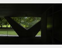 Dunkler überdachter Bereich mit einer Betonwand in V-Form, die ein großes Fenster zur Aussicht auf eine grüne Parklandschaft mit Bäumen und Rasenflächen einrahmt. Rechts sind leicht reflektierende Glasflächen sichtbar.