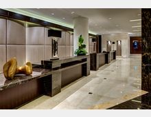 Elegante Hotellobby mit polierten Marmorböden, dunklen Marmortresen, einer beigen Wandverkleidung und moderner Dekoration, darunter Pflanzenarrangements, eine Skulptur und Designerlampen. Im Hintergrund sind Türen und ein Bildschirm erkennbar.