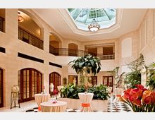 Großer, eleganter Innenraum mit heller Steinarchitektur, verziertem Geländer auf einer umlaufenden Galerie, Glasdach in achteckiger Form sowie schwarz-weißem Schachbrettboden. Im Zentrum stehen Pflanzenarrangements und dekorierte Stehtische.