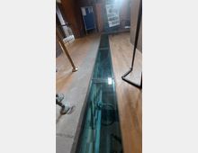 Innenansicht eines Museums oder Ausstellungsraums mit einem langen, schmalen Glasboden, der den Blick auf darunterliegende Kacheln und technische Installationen freigibt. Der Raum hat gemischte Bodenbeläge aus Holz und Fliesen sowie Ausstellungstafeln im Hintergrund.