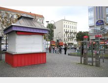 Ein kleiner geschlossener Verkaufskiosk mit roter Verkleidung und lilafarbener Dachleiste steht auf einem gepflasterten Platz. Im Hintergrund sind mehrstöckige Wohn- und Geschäftsgebäude, mehrere Passanten, Telefonzellen sowie eine Straßenkreuzung zu sehen.