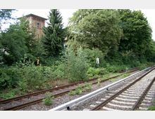 Bahngleise in einer grünen, bewachsenen Umgebung mit Bäumen und Sträuchern; im Hintergrund ist ein Backsteingebäude mit einem kleinen runden Fenster zu sehen. Ein Schild mit der Aufschrift „15/4“ steht links der Gleise.