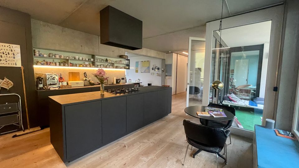 Moderne Wohnküche mit schwarzer Kücheninsel, Holzarbeitsplatte und eingelassenem Gasherd. Im Hintergrund Regale mit Geschirr, ein Esstisch mit Stühlen und eine verglaste Tür zu einem weiteren Wohnbereich.