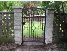 Ein schmiedeeisernes Gartentor mit dekorativen Ornamenten, eingefasst von grauen Steinsäulen und einer gemusterten Betonmauer, öffnet den Blick auf einen gepflegten Garten mit Rasen und Bäumen. Im Hintergrund ist ein Holzgebäude erkennbar.