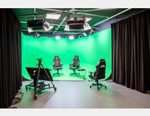 Ein moderner Studioaufnahmeraum mit grünem Greenscreen-Hintergrund, mehreren Studiostühlen, einem Kamerastativ und Scheinwerfern an der Decke. Schwarze Vorhänge rahmen den Raum ein, und Technikgeräte sowie Kabel sind am Boden sichtbar.