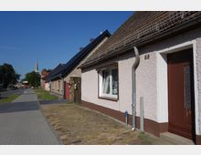 Straßenszene mit Reihenhäusern, deren Fassaden verputzt sind, und einem Bürgersteig aus Betonplatten. Im Hintergrund ist ein Kirchturm mit grünem Dach erkennbar, und im Vordergrund liegt ein gepflasterter Weg neben der Straße.