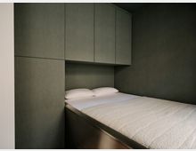 Schmaler Schlafbereich mit einem eingebauten Bett mit weißer Bettwäsche, umgeben von dunkelgrauen Wand- und Schrankpaneelen. Über dem Kopfteil befinden sich bündig integrierte Hängeschränke, das Bettgestell wirkt als Einbaumöbel mit Stauraum.