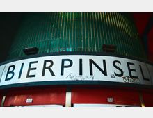 Nahaufnahme des Gebäudes "Bierpinsel" in Berlin, mit einem grünen, säulenförmigen Bereich mit vertikalen Linien und einem großen weißen Schild mit schwarzer Schrift, auf dem "BIERPINSEL" steht. Im unteren Bereich des Schildes sind Graffiti und Verschmutzungen sichtbar.