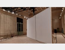 Großer Innenraum mit hohen Decken und einer modularen weißen Stellwand in der Mitte. Der Raum hat helle Wände, ein gemustertes Fliesenbodenmuster, Stahlträger an der Decke und eine grüne Doppeltür auf der linken Seite; Leitern und technische Geräte sind sichtbar.