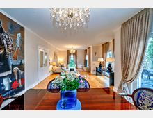 Elegantes Wohnzimmer mit großem Kronleuchter und bodenlangen Vorhängen an den Fenstern, eingerichtet mit einem Sofa, dekorativen Tischlampen und einem Flachbildfernseher. Im Vordergrund steht ein Esstisch mit einer Blumenvase, links eine auffällige Wandmalerei, und rechts führt eine Terrassentür ins Freie.