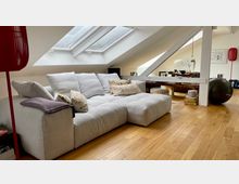 Wohnbereich in einem Dachgeschoss mit hellem Holzfußboden, einer großen, grauen Eckcouch mit dekorativen Kissen und schrägen Deckenfenstern. Im Hintergrund ein Essbereich mit Tisch und Stühlen, dazu ein schwarzer Gymnastikball und rote Lampen als Akzente.