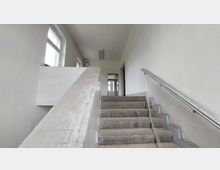 Ein leerer Treppenhausflur in einem schlichten Gebäude mit Betonstufen, einem Metallgeländer auf der rechten Seite und weißen, glatten Wänden. Durch die Fenster auf der linken Seite fällt Tageslicht herein.