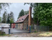 Kleines Feuerwehrgebäude mit rotem Ziegeldach, rundlichen Dachfenstern und einem hohen gemauerten Schornstein, gelegen hinter einem Metalltor. Im Hintergrund sind Bäume und ein grauer Himmel zu sehen.