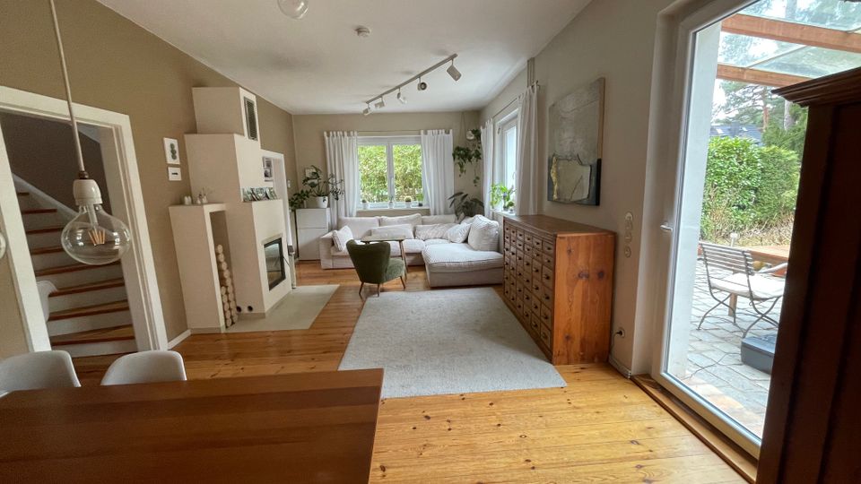 Helles Wohnzimmer mit Holzdielen, großem Ecksofa auf Teppich und einem Holzschrank mit vielen Schubladen an der Wand. Rechts führt eine bodentiefe Glasschiebetür auf eine Terrasse mit Tisch und Stühlen; links sind eine Treppe und ein moderner Kaminofen zu sehen.