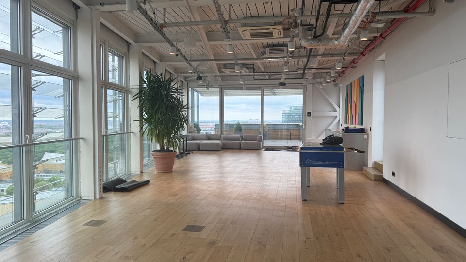 Open-plan loft interior with wood floors, floor-to-ceiling windows on two sides, and an exposed ceiling with visible ducts, pipes, and lighting. A potted tree, low sectional seating near the windows, and a foosball table sit in the spacious room with a city view beyond.