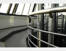 Innenansicht eines modernen Gebäudes mit einer gebogenen Glasfassade und einem Geländer aus Edelstahl. Der Boden ist aus Beton, und die Umgebung hat großzügige Fenster, die viel Licht hereinlassen.