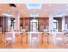 Großer Seminarraum mit sichtbaren Backsteinwänden, rechteckigen Tischen und weißen Stühlen auf Parkettboden. Großzügige Fenster mit Rundbögen lassen Tageslicht herein, ergänzt durch moderne Deckenspots und blaue Lichtakzente.