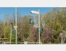 Eine Straßenecke mit Verkehrsschildern und Ampeln, darunter Wegweiser zu "Lichtenrade", "Ulsteinstraße" und "Rixdorfer Straße". Im Hintergrund sind blühende Bäume und dichte Vegetation zu sehen, unter einem klaren blauen Himmel.