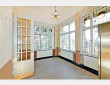 Heller Raum mit großen Buntglasfenstern, die detaillierte Muster zeigen, sowie einer weißen Tür mit Glaseinsatz. An der Decke hängt ein Kronleuchter, und unter den Fenstern sind Heizkörper angebracht.