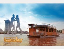 Hausboot auf der Spree in Berlin mit der Metallskulptur "Molecule Man" im Hintergrund; ein modernes Hochhaus und Uferbauten sind ebenfalls sichtbar unter einem bewölkten Himmel.