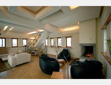 Moderner Wohnraum mit Holzfußboden, einem Kamin in der rechten Wand, Sitzsäcken und einem Sofa. Eine weiße Treppe mit schwarzem Geländer führt zur oberen Ebene, und mehrere Fenster lassen Tageslicht herein.