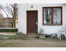 Ansicht eines Hauseingangs mit weißer Putzfassade, einer braunen Tür und einem angrenzenden Fenster mit Holzrahmen. Daneben stehen mehrere weiße Kunststoffstühle, und im Vordergrund ist ein kleiner gepflasterter Bereich mit ein paar Pflanzen in Betonkästen zu sehen. Im Hintergrund sind Wohnhäuser, Büsche und Bäume erkennbar.