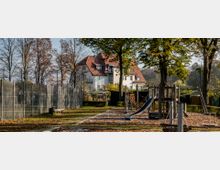 Spielplatz mit Holzklettergerüst, Rutsche und Hängebrücke, umgeben von Laubbäumen. Im Hintergrund steht ein großes weißes Gebäude mit roten Ziegeldächern, und links entlang des Platzes verläuft ein hoher Metallzaun.