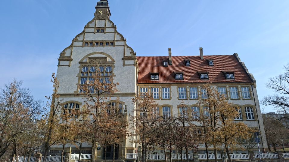 Historisches mehrstöckiges Gebäude mit heller Fassade, großen Sprossenfenstern und hohem geschwungenem Giebel mit Uhrturm, daneben ein langer Flügel mit rotem Ziegeldach und Gauben. Davor stehen mehrere laublose Bäume entlang eines Zauns unter blauem Himmel.