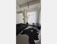 Heller, loftartiger Wohn- oder Arbeitsraum mit hohen Decken, weißen Backsteinwänden und großer Fensterfront mit transparenten Vorhängen. Im Vordergrund stehen ein schwarzes Ecksofa und ein schwarz-weißes Kuhfell auf Betonboden; daneben ein Topfbaum, ein kleiner Tisch mit Stuhl und ein blauer Hocker.