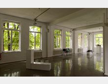 Ein heller Galerieraum mit blank poliertem Boden und großen, gewölbten Fenstern, die den Blick auf grüne Bäume freigeben. Im Raum stehen weiße Podeste mit Kunstobjekten sowie einige Stühle entlang der Wand.