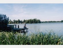 Ein See mit ruhiger Wasseroberfläche, umgeben von Bäumen am Ufer. Im Vordergrund sitzt eine Person auf einem hölzernen Steg neben Schilf.