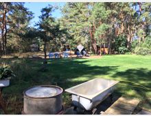 Großer Garten mit Rasenfläche, umgeben von hohen Kiefern und anderen Bäumen. Im Vordergrund stehen eine alte Badewanne und ein Metallbehälter, während im Hintergrund Plastikstühle und Liegen, teils unter einem Sonnenschutz, stehen.
