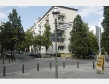 Straßenszene am Karl-August-Platz in Berlin mit mehrstöckigen Wohnhäusern im Hintergrund, umgeben von Bäumen. Im Vordergrund befindet sich ein Straßenlaternenmast mit Straßenschild und ein Wahlplakat mit der Aufschrift „Berlin kann mehr.“