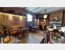 Gemütlicher Innenraum eines Cafés oder Restaurants mit Backsteinwänden, Holzboden und antikem Mobiliar. Mehrere Esstische mit gepolsterten Stühlen sind verteilt, an einer Wand steht ein Regal mit Flaschen und Gläsern, und eine warme Beleuchtung schafft eine einladende Atmosphäre.