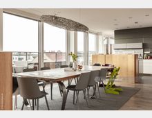 Offener Wohn- und Essbereich mit einem großen Holzesstisch und mehreren Stühlen, umgeben von bodentiefen Fenstern, die auf eine Terrasse mit Blick auf die Dächer einer Stadt führen. Im Hintergrund befindet sich eine moderne Küche mit weißen Schränken und einer Trennwand aus Holz.