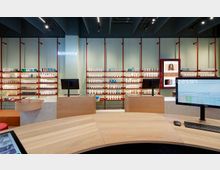 Innenraum einer modernen Apotheke oder eines Kosmetikgeschäfts mit hellen Regalen, die verschiedene Produkte ausstellen. Im Vordergrund befinden sich hölzerne Verkaufstheken mit Computermonitoren, und im Hintergrund hängt ein Neon-Schriftzug mit dem Wort "kosmetik".