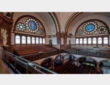 Innenraum einer historischen Kirche mit roten Backsteinwänden, hohen Gewölbedecken und farbigen Rosettenfenstern. Holzbänke sind auf zwei Ebenen angeordnet, und dekorative Arkaden und Statuen schmücken den Raum.