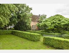 Ein gepflegter Garten mit symmetrischen Buchsbaumhecken, Rasenflächen und dicht belaubten Bäumen; im Hintergrund befindet sich ein rot geklinkertes Gebäude mit einem großen Holztor.
