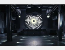 Dunkler Innenraum eines Clubs oder Veranstaltungsortes mit zentraler Bühne, über der eine kreisförmige LED-Beleuchtung mit strahlenförmigem Muster angebracht ist. Industriearchitektur mit freiliegenden Balken und stimmungsvoller Beleuchtung auf dem Boden.