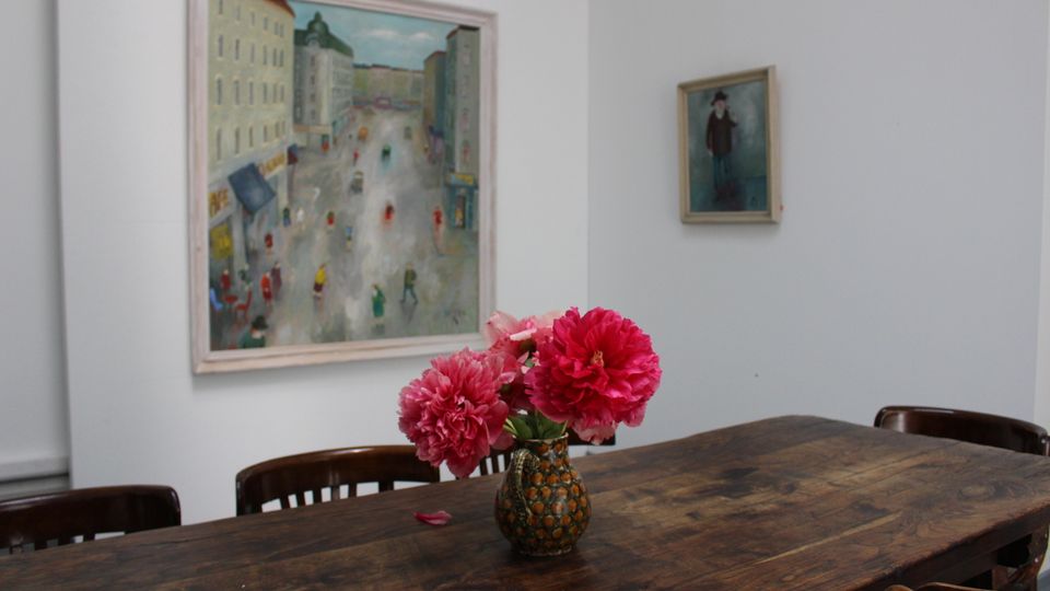 Ein heller Innenraum mit weißer Wand, an der zwei gerahmte Gemälde hängen. Im Vordergrund steht ein langer, dunkler Holztisch mit Stühlen und einer Vase mit pinken Blumen in der Mitte.