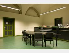 Innenraum eines Büros mit gewölbter Decke, grünem Boden und einer grünen Tür. Im Vordergrund stehen schwarze Tische mit Stühlen, im Hintergrund befinden sich eine dunkle Empfangstheke und Regale mit Akten.