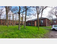 Backsteingebäude mit großen Fenstern in einem parkähnlichen Areal, umgeben von Bäumen und grüner Wiese. Ein kleiner, weißer PKW steht rechts auf einem unbefestigten Weg.