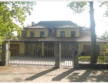 Zweistöckiges Einfamilienhaus mit gelber Fassade und dunklem Ziegeldach, umgeben von einem Garten. Vor dem Haus steht ein geschmiedetes Metalltor mit zwei steinernen Säulen.